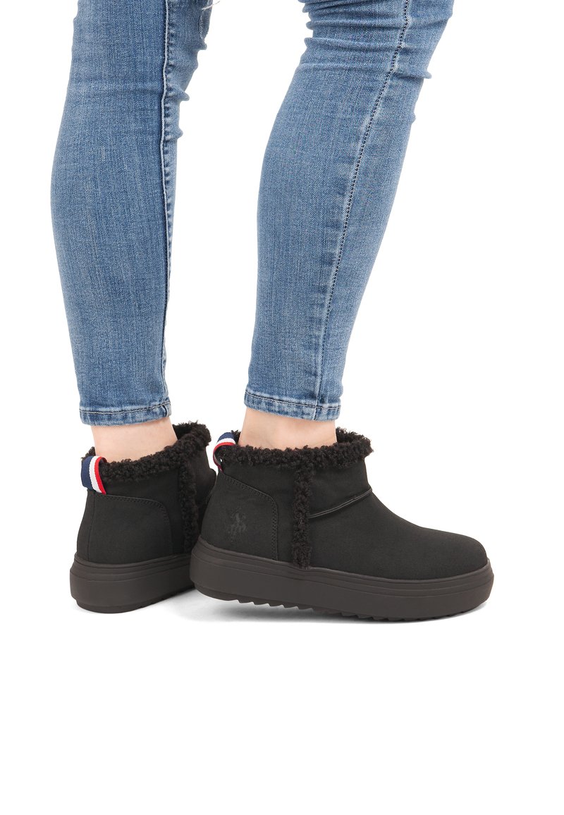 Person, der schwarze, fleecegefütterte Stiefeletten mit dicken Sohlen und Jeans trägt, steht vor einem weißen Hintergrund.