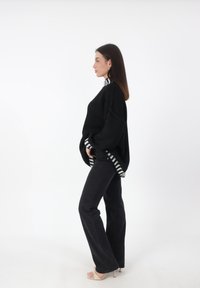 Zwarte oversized gebreide trui met een gestreepte halslijn, gecombineerd met zwarte flared jeans en lichtgekleurde sandalen met bandjes, bekeken vanaf de zijkant.