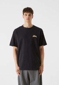 Melns kokvilnas T-krekls ar īsām piedurknēm un apaļu kakla izgriezumu. Zelta logo akcenti kreisajā krūtīs, brīvs piegriezums, gluda tekstūra.