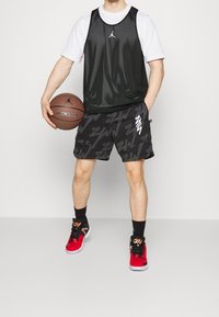 Svart nät-baskettröja över en vit t-shirt, tillsammans med mönstrade svarta shorts. Röda och svarta sneakers; håller en brun basketboll.