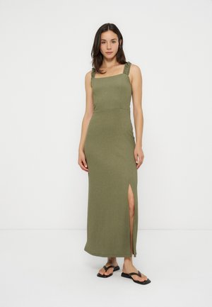 Femme en robe sans manches vert olive avec bretelles volantées et une fente haute sur la cuisse, portant des sandales plates noires, debout devant un fond blanc.