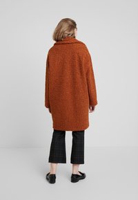 Orange fluffig kappa med hög krage, långa ärmar och en oversized passform, ihop med svarta rutiga cropped-byxor och svarta skor.