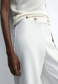 Witte op maat gemaakte broeken met een gladde textuur, voorzien van gouden decoratieve knopen op de tailleband. Zacht crèmekleurig sweater gedeeltelijk zichtbaar.