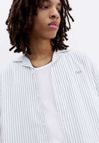 Bílá pruhovaná košile s zapínáním na knoflíky a límečkem ve stylu kempu, zdobená světle modrým vyšíváním. Nositelná přes bílý texturovaný tílko.