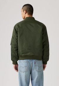 Mann med kort, mørkt hår iført olivengrønn bomberjakke og lyseblå jeans, stående med ryggen mot en ensfarget bakgrunn.