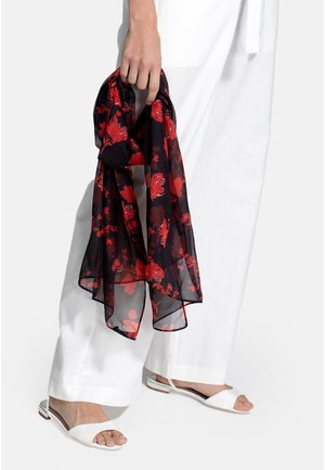 Hand holding a black sheer scarf with red floral print, wearing white wide-leg pants and white open-toe flat sandals.
