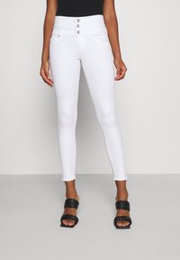 Jeans blancos ajustados con una cintura alta, con tres botones frontales y un acabado suave. Combinados con sandalias negras de tiras.
