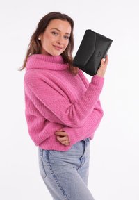 Černá syntetická kabelka na ruku s texturovaným povrchem, úhlovým designem a výrazným logem. Držená v ruce vedle růžového pleteného svetru.