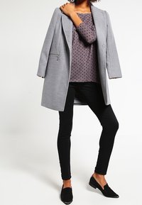 Grau melierter Wollmix-Mantel mit taillierter Passform, langen Ärmeln und seitlichem Reißverschluss. Kombiniert mit einem lila gepunkteten Oberteil und schwarzen Skinny-Jeans. Schwarze Loafers.