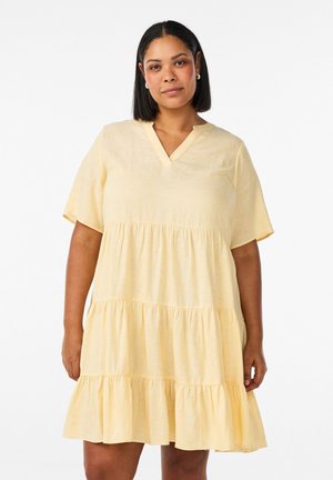 Žena so stredne dlhými čiernymi vlasmi, oblečená v krátkymi rukávmi a svetložltých vrstvených šatách, stojaca pred jednoduchým bielym pozadím.