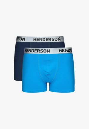 Oberkörperfreier Mann, der mit einer Hand hinter dem Rücken steht und leuchtend blaue Boxershorts mit einem silbernen Bund trägt, auf dem "HENDERSON" steht.