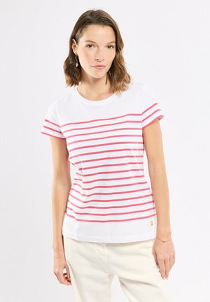 Jeune femme portant un t-shirt blanc à rayures horizontales roses et un pantalon clair, debout devant un fond uni.