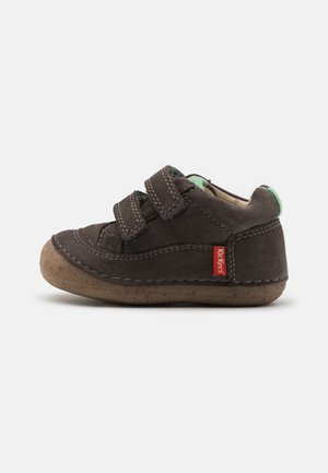 Zapato infantil de ante marrón con dos correas de Velcro, punta redonda, suela de goma y acento verde en el talón. Presenta costuras decorativas y un logo rojo.