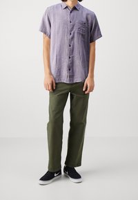 Camisa de manga corta con botones en color púrpura claro con un patrón sutil, combinada con pantalones verde oliva y zapatillas negras con suelas blancas.