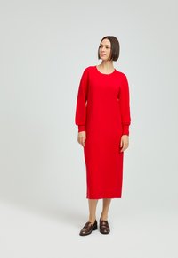 Rotes Kleid mit langen Ärmeln, Rundhalsausschnitt und gerader Silhouette. Glatte Textur, knielang und gerippte Bündchen. Braune Schuhe.
