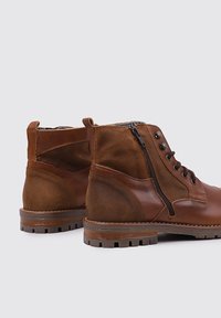 Bottes en cuir marron avec une finition en daim, dotées d'œillets en métal, d'une fermeture éclair sur le côté et d'une semelle en caoutchouc texturée.