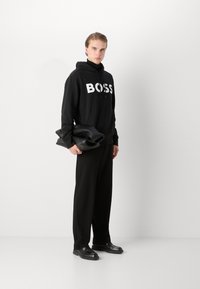 Juodas gobtuvas su didele balta "BOSS" logo, derinamas su laisvomis juodomis kelnėmis ir juodais batais, laikantis juodą odinį krepšį.