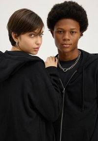 Kaksi nuorta aikuista, jotka pukeutuvat mustiin hupullisiin paitoihin; toisella on lyhyet ruskeat hiukset ja rengaskorvakorut, toisella afro-tyyli ja hopeinen ketjuriipus.