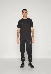 Vyras, vilkintis juodą Puma King marškinėlį ir derančias juodas Puma King kelnes bei juodais sportiniais bateliais, prieš paprastą šviesią fono.