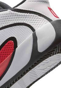 Primer plano de un zapato deportivo blanco, negro y rojo que muestra una parte superior de malla texturizada, curvas negras y suela con patrón.