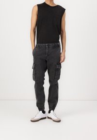 Černý top bez rukávů a tmavě šedé kalhoty typu cargo s elastickými manžetami. Kalhoty mají boční kapsy a volný střih. Celý vzhled doplňují bílé tenisky.