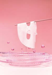 Biała odzież z dużymi wycięciami wisi na różowej rurze nad przezroczystymi, wzorzystymi talerzami i różowymi kulami, na miękkim różowym tle.