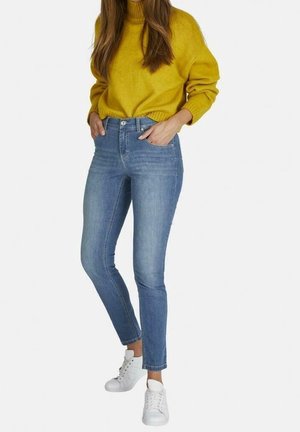 Mujer con un suéter amarillo mostaza, jeans ajustados azul claro y zapatillas blancas, de pie con las manos en los bolsillos frente a un fondo blanco.