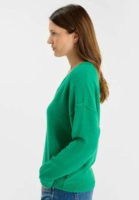 Groene gebreide trui met een V-hals, lange mouwen en zijsplitten aan de onderrand. De textuur lijkt zacht en iets viltachtig.