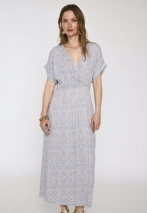 Femme portant une robe longue bleu clair à motifs, manches courtes et col en V, debout sur un fond blanc uni.
