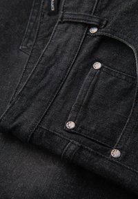 Prenda de mezclilla negra con costuras visibles, remaches grabados con el nombre de la marca y detalles de costura en el pequeño bolsillo delantero.