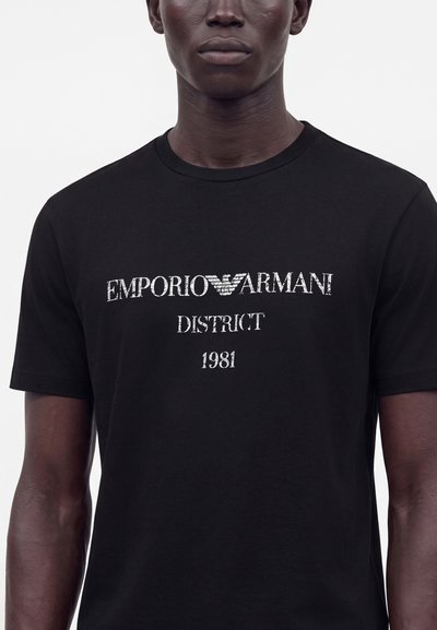 Άνδρας που φοράει μαύρο μπλουζάκι με λευκό κείμενο "EMPORIO ARMANI DISTRICT 1981" και λογότυπο αετού μεταξύ των λέξεων.