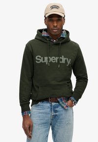 Mørkegrøn hættejakke med stort gråt "Superdry" logo, lagdelt over en ternet skjorte. Båret med lyseblå jeans og et brunt bælte. Kasket på hovedet.