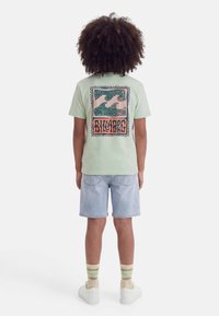 Göndör hajú gyerek világoszöld pólóban, amelyen hullám és szöveg minta található a hátulján, világos farmer rövidnadrágban, csíkos zokniban és fehér cipőben.