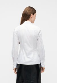 Γυναίκα με μακριά καστανά μαλλιά, φορώντας εφαρμοστό λευκό πουκάμισο με κουμπιά και μαύρη δερμάτινη φούστα, που βλέπει από πίσω σε απλό φόντο.