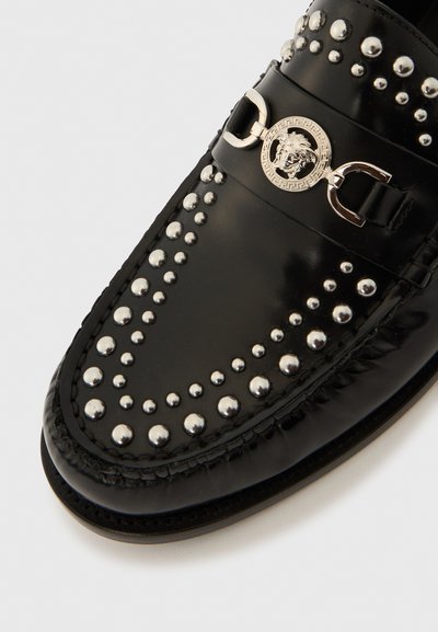 Mocasini negri din piele, cu ținte argintii, o cataramă decorativă și un emblema cu leu centrată pe bretea. Textură netedă și formă rotunjită.