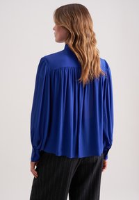 Blusa di seta blu con maniche lunghe, dettagli arricciati sulle spalle e vestibilità ampia. Tessuto liscio, abbinata a pantaloni scuri a righe.