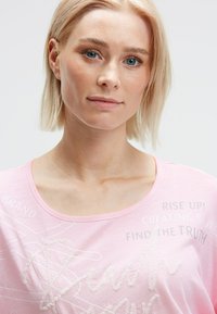 Blonde woman with short hair and blue eyes wearing a light pink top with white text and sequin details stands against a plain background.