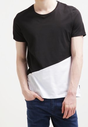 Persoon draagt een zwart-wit T-shirt met korte mouwen in kleurvlakken en een blauwe spijkerbroek, staand met één hand in de zak.