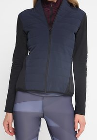 Veste matelassée bleu marine à col montant et fermeture éclair complète, associée à des manches longues noires. Leggings gris marbré avec des motifs angulaires.