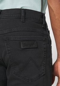Los jeans de mezclilla negra presentan un bolsillo trasero con un diseño de "W" cosido y una etiqueta rectangular estampada con "Wrangler". Se aprecia una tela texturizada.