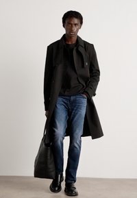 Abrigo largo negro sobre una camisa negra, jeans azules y zapatos negros. Sosteniendo una bolsa negra, de pie contra un fondo neutro.