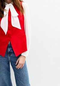 Rote Weste mit V-Ausschnitt und fünf Knöpfen, kombiniert mit einer weißen Bluse mit Schleife. Dazu weite blaue Jeans.