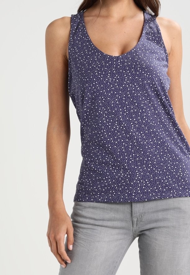 Femme portant un haut sans manches bleu marine avec des points blancs en forme d’étoiles et un pantalon gris clair ajusté, debout devant un fond uni clair.
