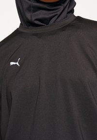 Svart idrottströja med en åtsittande design, tillverkad av ett mjukt, elastiskt tyg. Har en liten vit Puma-logotyp på vänster sida.