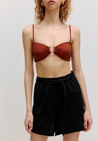 Bruine bikini top met geplooide textuur, met een ronde gouden accent in het midden; gecombineerd met losse zwarte shorts en een gestrikt taille-detail.