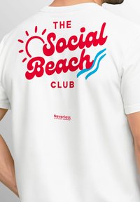 Weißes T-Shirt mit rotem und blauem Text: "DER Social Beach CLUB", mit Sonnen- und Wellen- Grafiken. Enthält das "Neverless"-Logo in rot.