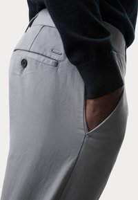 Pantaloni grigi chiari realizzati in tessuto liscio, con una tasca frontale, una tasca sul retro con bottone e un'etichetta del marchio discreta vicino alla vita.
