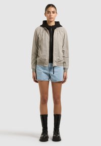 Jeune femme portant une veste beige à fermeture éclair par-dessus un sweat à capuche noir, un short en jean bleu clair et des bottes noires à lacets, debout devant un fond uni.