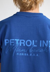 Modré bavlněné tričko s výrazným potiskem na zadní straně, který zahrnuje "PETROL IND," "Miami beach" a "FLORIDA, U.S.A." v různých odstínech modré.