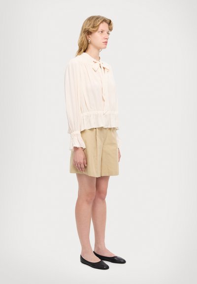 Blouse beige clair avec un col à nœud et une taille froncée, associée à un short beige. Le modèle porte des ballerines noires. Texture de tissu lisse.